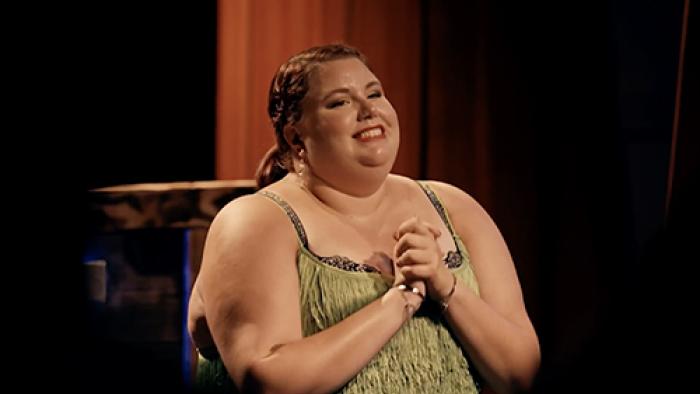 Thick Skin TV show screenshot 3: A smiling brunette woman in a green fringed dress clasps her hands at her chest and smiles.
