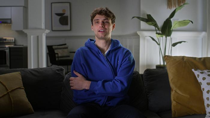 Thirst Trap TV show screenshot 1: William White sits on a dark grey couch. He wears a royal blue sweatshirt and smiles with his arms crossed