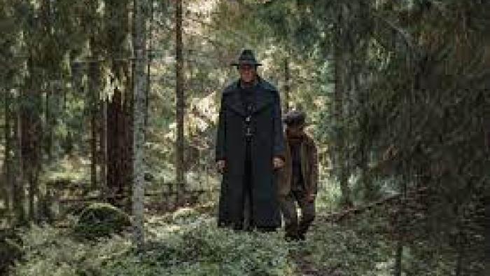 To Cook a Bear TV show screenshot 3: Two men, one tall, and one shorter, walk through a dense forest wearing dark clothing atop a forest floor of moss