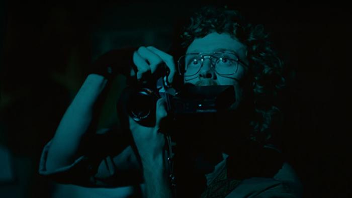 True Haunting TV show screenshot 3: A man with curly hair and 1980s-style metal aviator glasses holds a camera up in front of his face