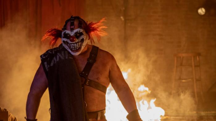Twisted Metal: Sweet Tooth is shown from the waist up, silhouetted against a roaring fire. His hair is wild and red and he wears a menacing clown mask.
