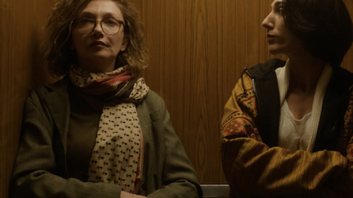 Unconditional TV show screenshot 3: Two people in an elevator, one in glasses and scarf, the other in a patterned jacket.