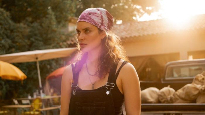 Under a Dark Sun TV show screenshot 1: A younger woman in black overalls, tanktop and a pink bandana stands looking, focused and offscreen
