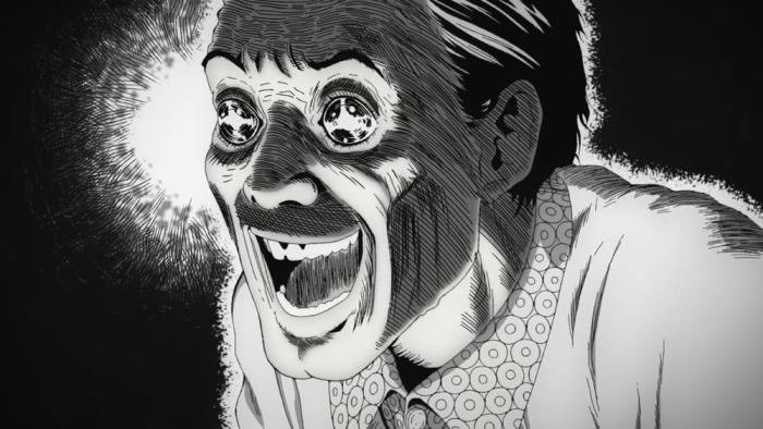 Uzumaki TV show screenshot 2: A man with spirals in his eyes grins widely. Black background with burst of white near his face to emphasize eyes.