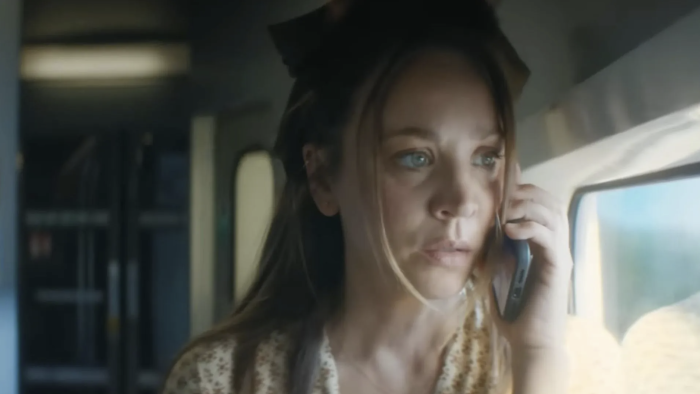 Vanished TV show screenshot 3: Cuoco holds a phone in her hands. She stands in a train cart. She wears a print dress and green ribbon in her hair.