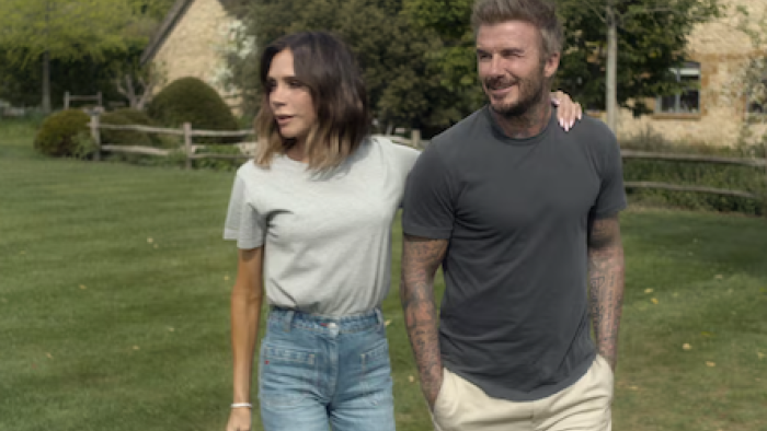 Victoria Beckham TV show screenshot 3: Victoria Beckham in gray shirt walks with David Beckham in blue shirt.
