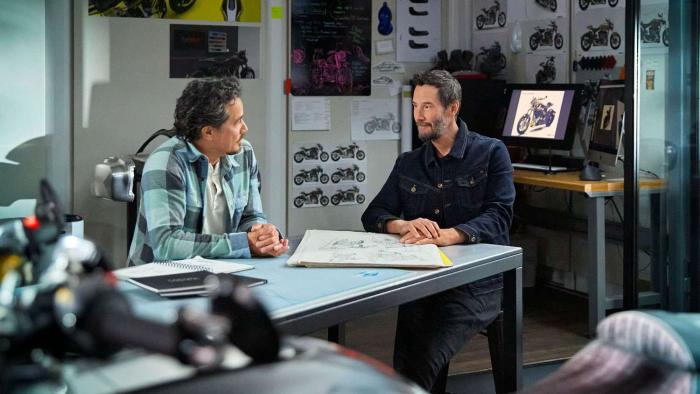 Gard Hollinger and Keanu Reeves sit at a table in a workshop. They are surrounded by motorcycle posters and images.