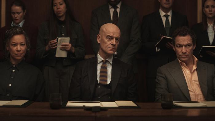 War TV show screenshot: Three lawyers seated at a table during a formal meeting, with others standing behind.