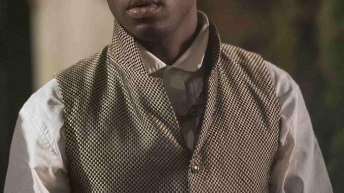 Ernest Kingsley Junior wears a checker patterned waistcoat over a white dress shirt. A gold chain hangs off one of the buttons.