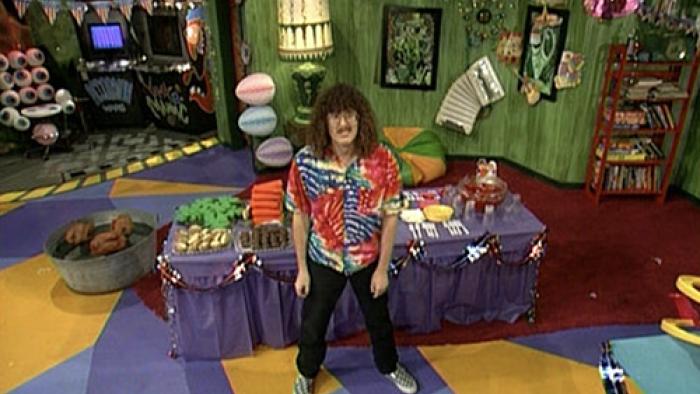 Weird Al stands in the middle of his colorful cave/clubhouse, looking up at the camera with a slight grimace.