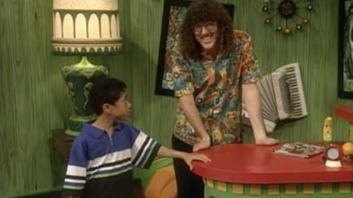 Weird Al stands at a green-and-red counter, chatting with a boy in a blue and black shirt.