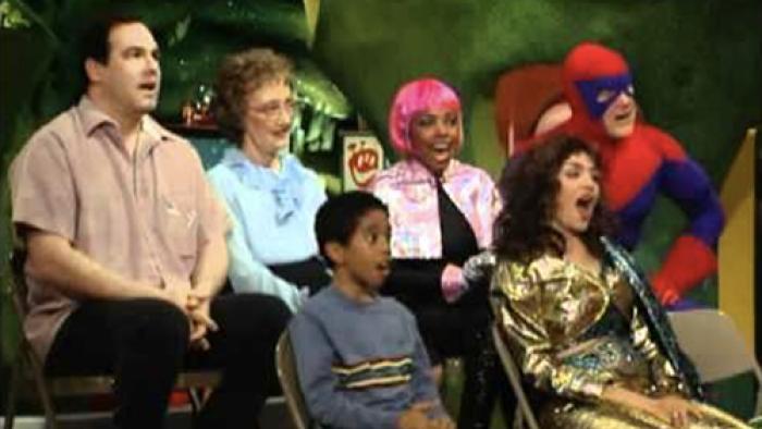 A small audience made up of kids, superheroes, gold lamé-clad comediennes, and others with expressions of amazement on their faces.