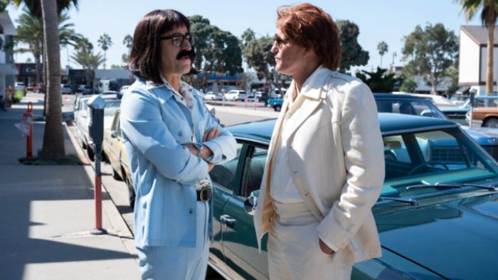 White House Plumbers TV show: Liddy and Hunt, in 1960s suits, stand in front of a vintage car on a street. Liddy wears a powder blue suit and black Prince Valiant-style wig; Hunt wears a beige suit and a rumpled orange wig
