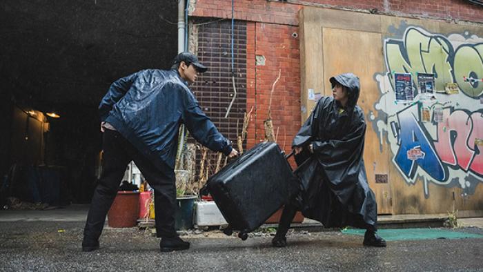 The Wonderfools TV show screenshot 2: A man and a woman in rain ponchos struggle over a black rolling suitcase in a gritty alley.