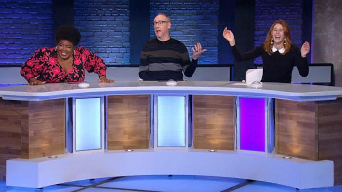 Comedians Dulcé Sloan, Matt Walsh, and Hannah Pilkes are seated at a long paneled desk on a game show set. Each person has a buzzer in front of them. Sloan and Pilkes are animatedly laughing, while Walsh appears to be speaking.