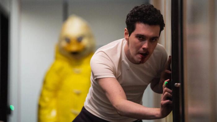 Wreck TV show: James stands in front of a cabin door in a t-shirt, it appears that he's attempting to listen in on what's inside. Behind him, a person in a yellow duck costume stands ominously. 