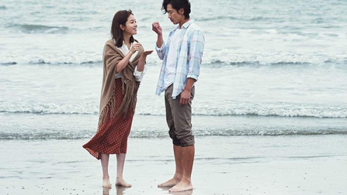 A man and woman stand barefoot on a misty-looking beach. The woman smiles and wears a tan blanket around her shoulders.