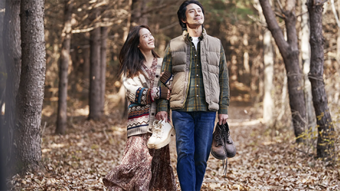 A man and woman walking through a forest in autumnal clothing, holding their shoes. The woman holds the man's arm, and they both have beatific expressions.