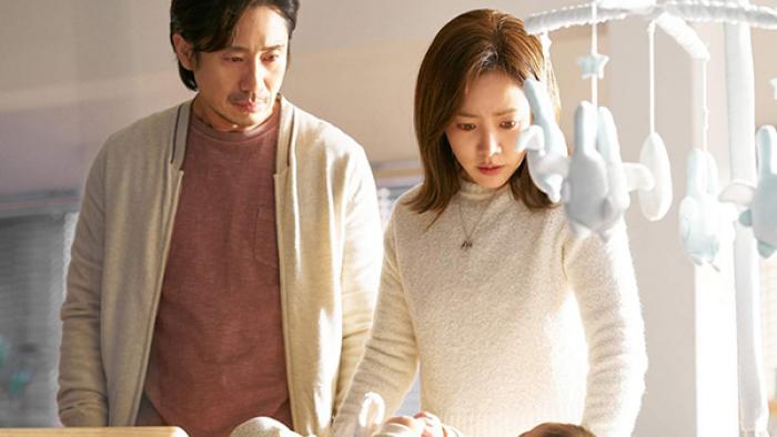 A man and woman stand over a crib with a mobile hanging over it. They look down at a baby, his expression somewhat blank and hers a bit unnerved.