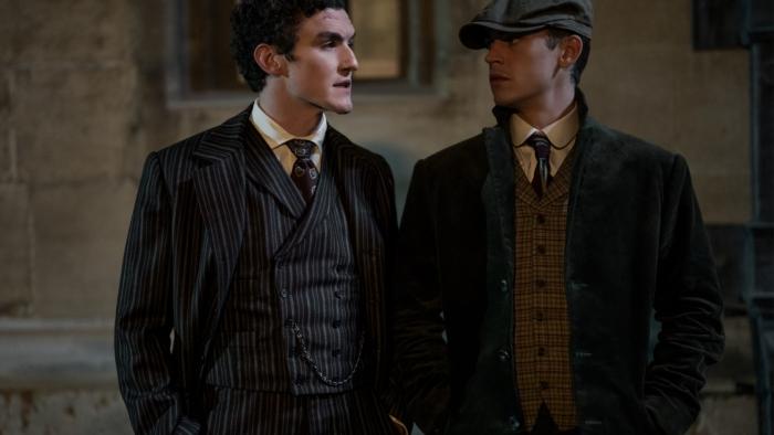 Young Sherlock TV show: Dónal Finn and Hero Fiennes Tiffin. Finn wears a striped suit. Tiffin wears a cap, striped vest, and a dark coat.