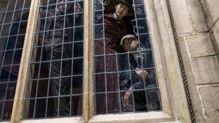 Young Sherlock TV show: Dónal Finn, Zine Tseng, and Hero Fiennes Tiffin. They stand against a broken window. Tseng and Tiffin