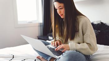Young teen in a sweater sitting on her bed working on a laptop.