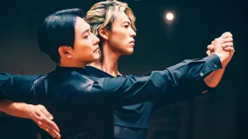 10Dance movie: Two Japanese men wearing black attire, left with black hair, right center with blond hair, clasp hands and dance together in a studio