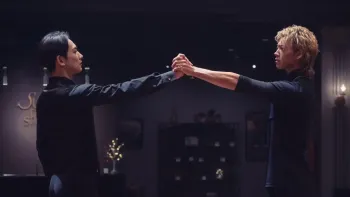 10Dance movie: In a dark dance studio, Japanese man with short black hair, left, reaches out and clasps hands with Japanese man with blond hair, right