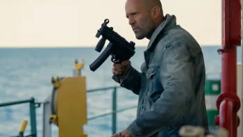 Mutiny: Shaved-headed man in denim jacket holding a gun on a boat, ocean in the background
