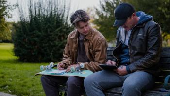 5lbs of Pressure Movie: Jimmy (Rudy Pankow) holds his upside-down skateboard on his lap, taking a drawing lesson from Adam (Luke Evans)