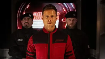 The Running Man: Glen Powell in red and black uniform stands with two security personnel behind him