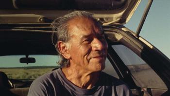 A Love Song: A close-up of a Native American man in his 60s sitting in the open trunk of his car