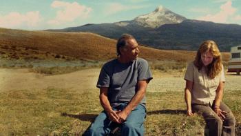 A Love Song: A Native American man and a blond White woman, both in their 60s, sit together on the grass, with mountains in the background