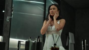 A Normal Woman: Milla in a nightgown stands in front of a large bathroom mirror touching her cheeks while looking at her reflection.