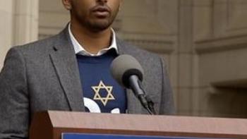 A Tree of Life movie: Wasi Mohamed, a South Asian American and former head of the Islamic Center of Pittsburgh, wearing a blue pullover top with a Star of David, white shirt, and gray jacket while speaking at a podium