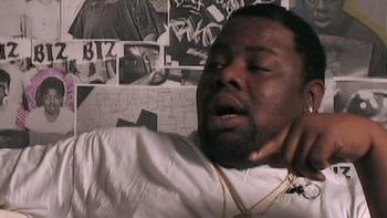 Still from All Up in the Biz: Biz Markie speaking while seated in front of a collage of clips about him, wearing a white T-shirt and gold chain necklaces 