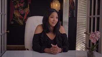 Still from All Up in the Biz: Tara Hall, the wife of Biz Markie, with a graphic illustration of her husband in the background, seated with hands folded together while in a white chair in front of a white desk, wearing a black V-neck halter top with cutout shoulders and long sleeves