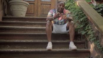Still from All Up in the Biz: Big Daddy Kane duo dancer Johnny Famous a.k.a. Scoop Lover speaking while seated on the brownstone stoop, wearing designer glasses, a watch, a ring, white shorts, white sneakers, and a multicolored top 