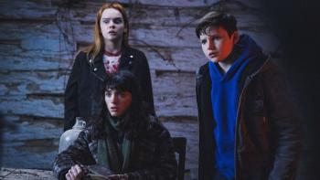 All Fun and Games Movie: In an old cabin, Billie (Natalia Dyer), who's seated; Sophie (Laurel Marsden), who's behind her; and Jo (Benjamin Evan Ainsworth), who's to the right, stare in horror at something spooky