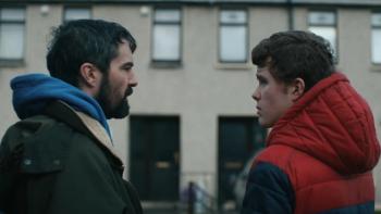 Amongst the Wolves: A bearded middle-aged White man talks to a munch younger White man outside a building 