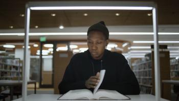 As We Speak: Rap Music on Trial movie: Rapper Kemba, a Black man, in black hoodie and hat sits reading a book in a library