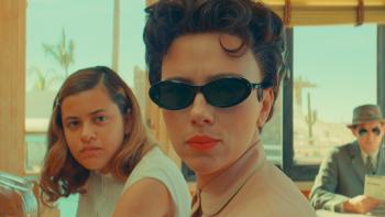 Asteroid City Movie: A woman in the 1950s wearing black sunglasses and red lipstick gives a hard look while sitting next to a teen girl