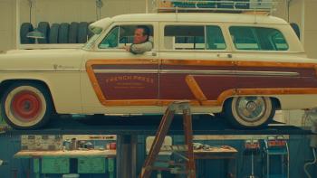 Asteroid City Movie: A middle-aged White mechanic is sitting inside a "Woody" wagon on a car hoist while two men look up at him