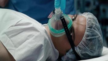 Lena Olin lays on her side in a hospital gown with a gas mask strapped to her face and a hairnet.