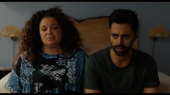 Babes Movie: Michelle Buteau and Hasan Minaj sit next to each other on a bed, looking distressed