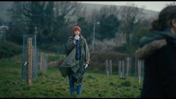 Bad Apples: White woman wears a warm hat and coat as she walks through a field sipping from a cup