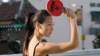 A close-up of Maggie Q as she holds a ping pong racket up on a sunny rooftop. Her hair is tied back in a ponytail and she is wearing a spaghetti strap top.