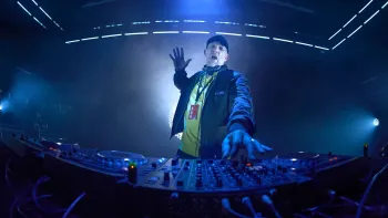 Banger movie: White French man in baseball hat, jacket, center, DJs and dances in front of turntables and equipment, dark lights all around him 