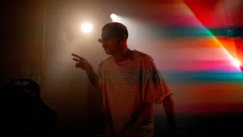 Banger movie: Black French man with short dyed blond hair wears long t-shirt, sunglasses, and a neck chain center in colorfully lit dark room
