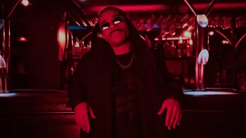 Banger movie: Black French man in black hoodie shawl, chain, and sunglasses, center, leans back against a bar with bottles/glassware in a red-lit club